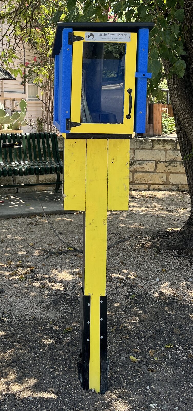 A colorful yellow and blue Little Free Library box shaded by a tree
