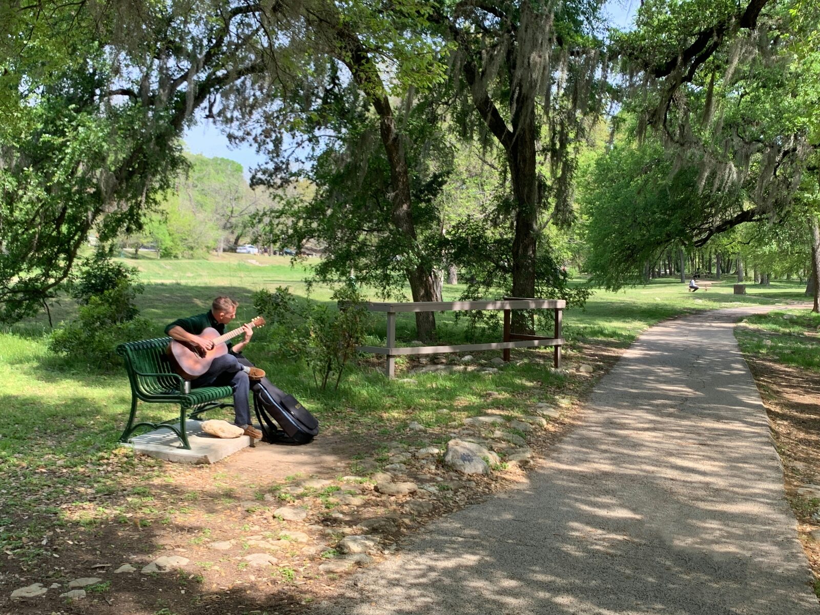 A musician plays guitar on a bench along a shaded Austin trail